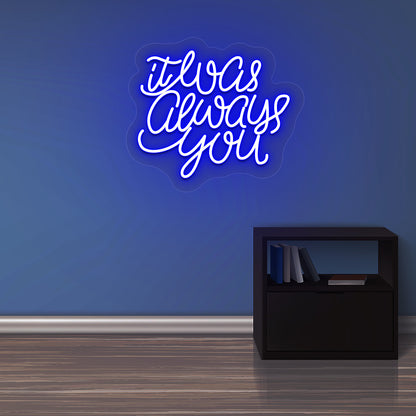 An image of an "It was always you" LED neon sign glowing on a wall in a warm blue color. 