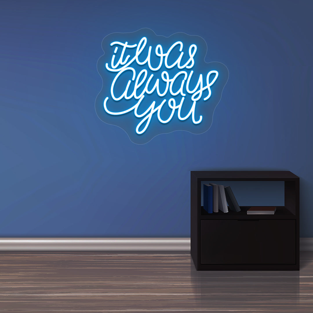 An image of an "It was always you" LED neon sign glowing on a wall in a ice blue color. 