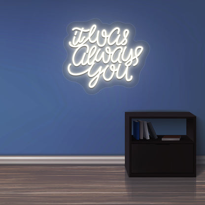 An image of an "It was always you" LED neon sign glowing on a wall in a warm white color. 