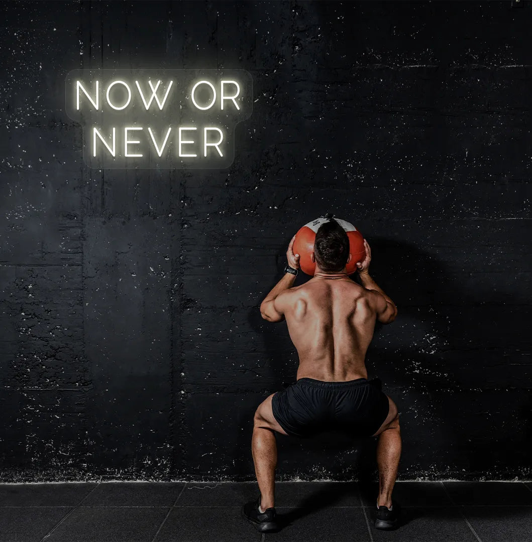 Trendy "now or never" neon sign in warm white mounted on fitness wall.