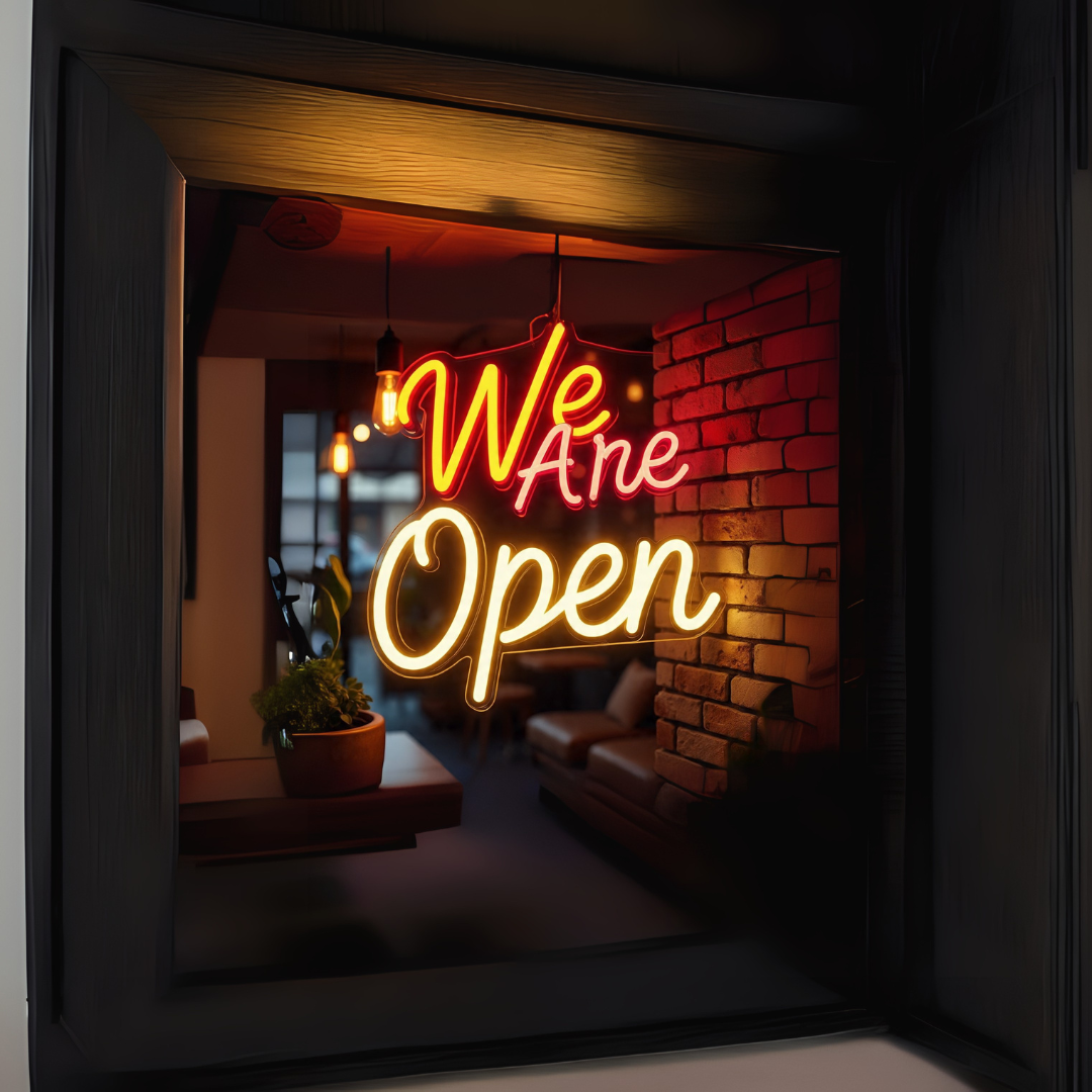 Image of a "We are open" LED neon sign in orange, red, and warm white colors on storefront glass wall.