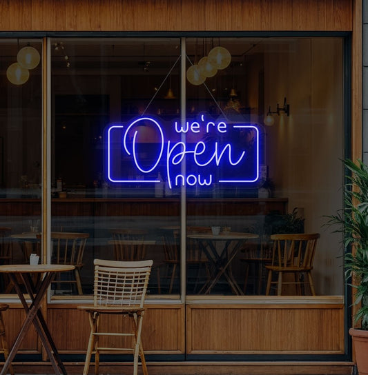 Image of a "We're open now" neon sign glowing on a cafe glass door in blue color.
