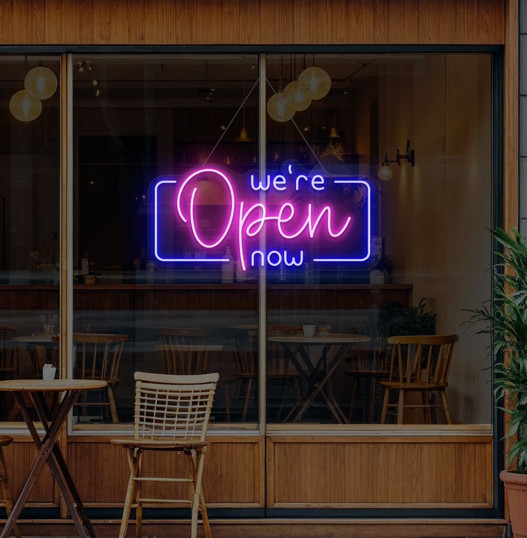 Image of a "We're open now" neon sign glowing on a cafe glass door in pink color.
