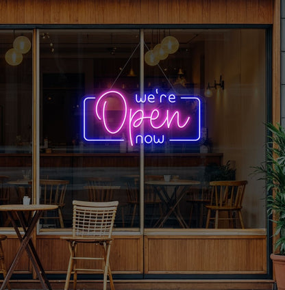 Image of a "We're open now" neon sign glowing on a cafe glass door in pink color.