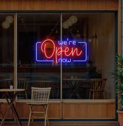 Image of a "We're open now" neon sign glowing on a cafe glass door in red color.