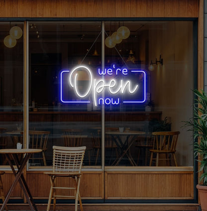 Image of a "We're open now" neon sign glowing on a cafe glass door in white color.