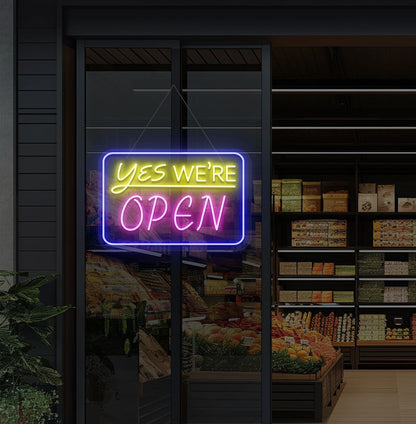 Image of a "Yes, we're open" neon sign glowing on a cafe glass door in pink color.